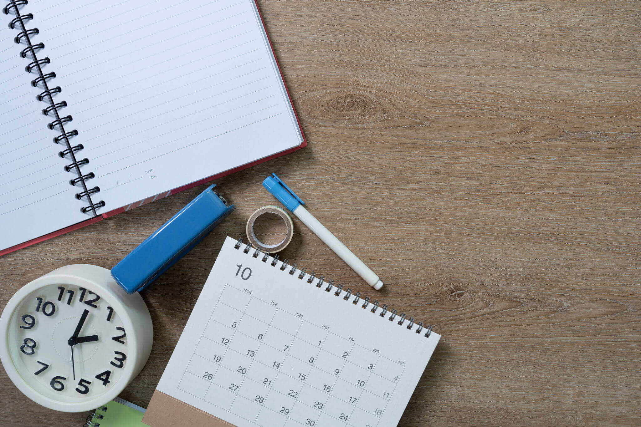 A clock, a calendar, an open notebook, a blue stapler, a pen, and tape are arranged on a wooden desk.