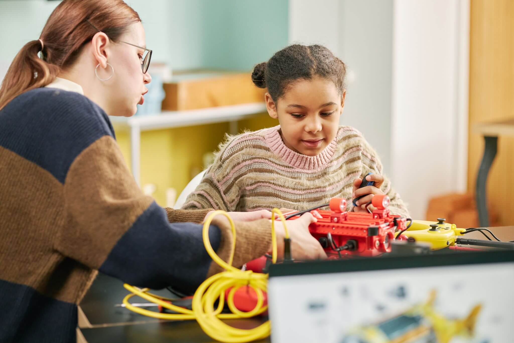 An adult and a child sit at a table working together on an electronics project with wires and devices in a classroom or workshop setting.