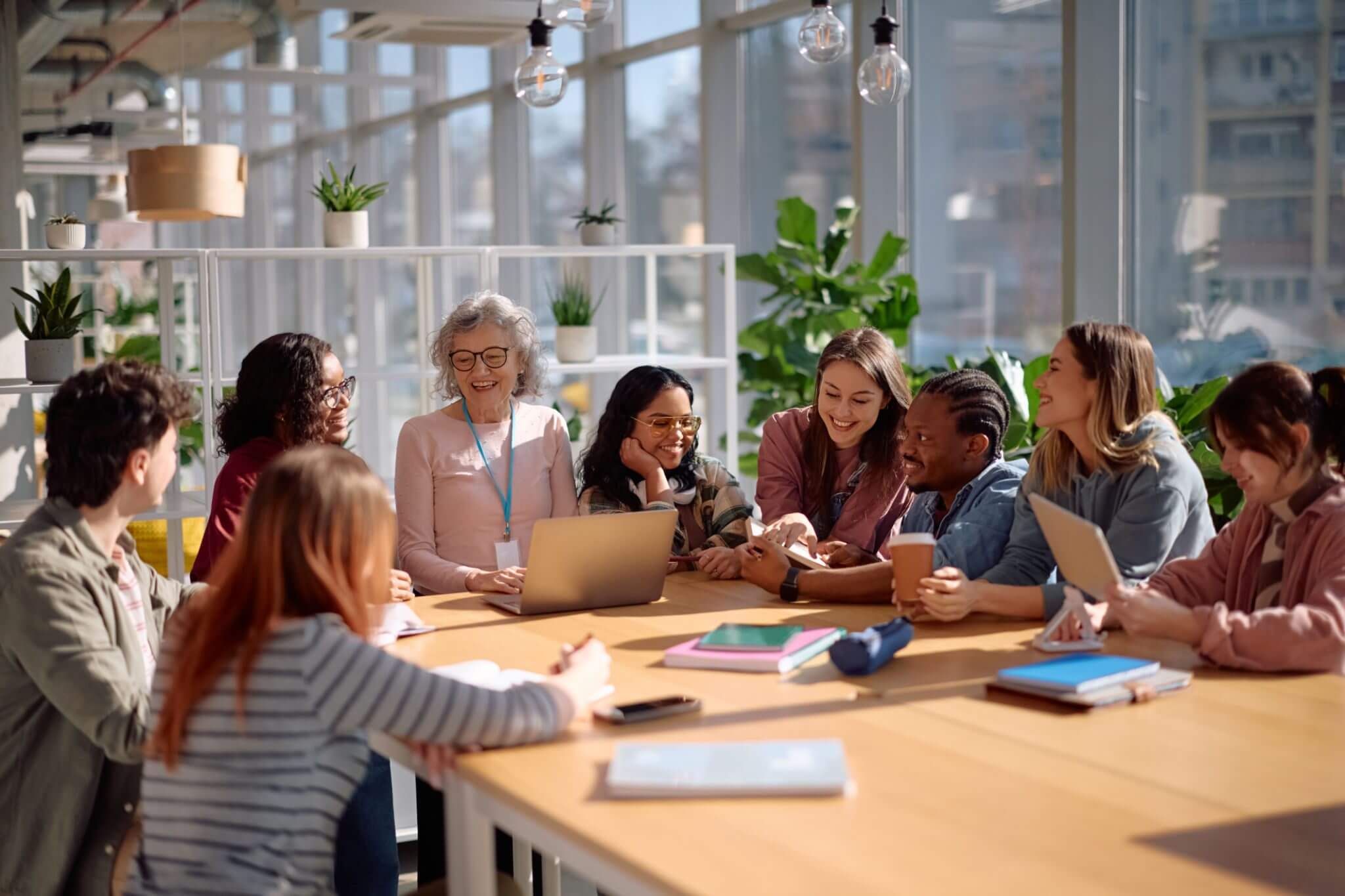 A group of people gather around a table in a bright room, engaged in conversation and smiling, with notebooks, laptops, and plants visible on the table and shelves.