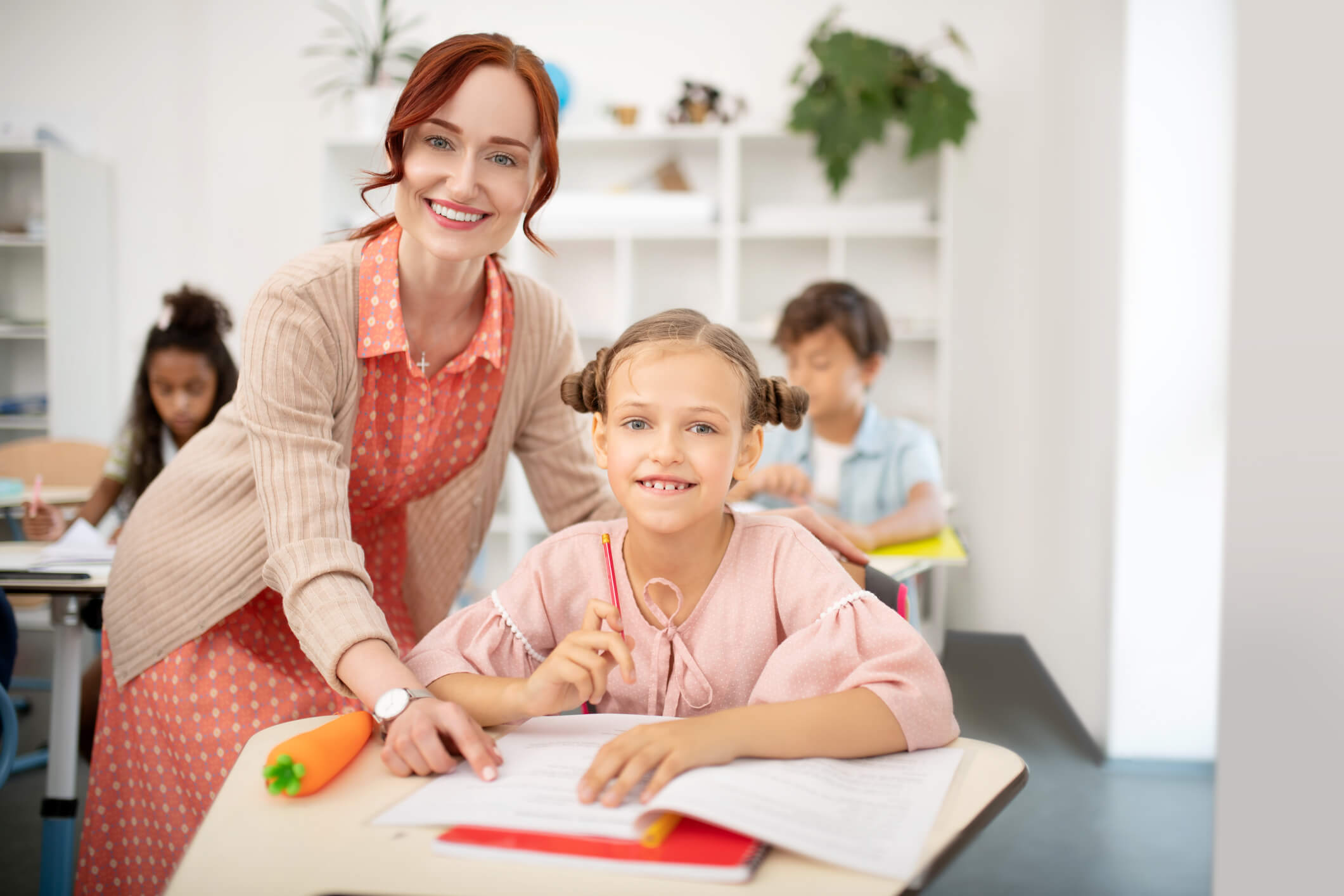 A teacher and a student smile at the camera while working together at a desk in a classroom, with other students visible in the background.