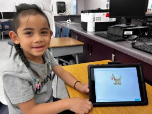 A young girl sits at a desk in a classroom, smiling, with a tablet displaying an educational image of a cartoon dog holding a red ball.