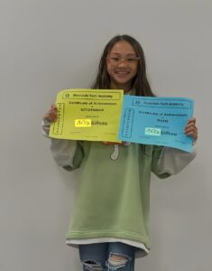 A person wearing glasses and a green sweatshirt holds two certificates for Citizenship and Math achievement from Riverdale Tech Academy.