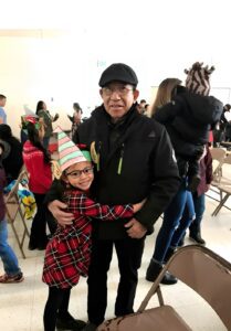 An older man in a black jacket and cap hugs a young girl wearing a holiday hat and plaid dress. They stand in a room with other people and folding chairs.